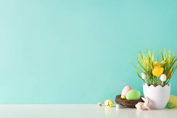 istock Easter decor idea. Side view shot of a table showcasing a shell-inspired vase with grass and tulips, a ceramic rabbit, and nest with vibrant eggs against a soft turquoise backdrop for text 2047995772