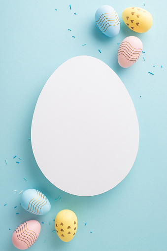 Charming Easter scene: vertical top view of multicolored eggs and sprinkles on a pastel blue surface, complemented by bunny ears behind an oval card for special notes or adverts