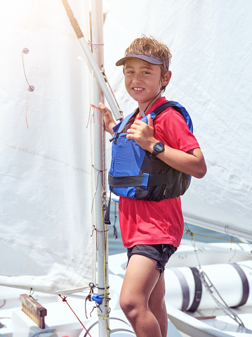 Positive male child in safety vest and cap smiling and looking at camera while standing on yacht during summer vacation
