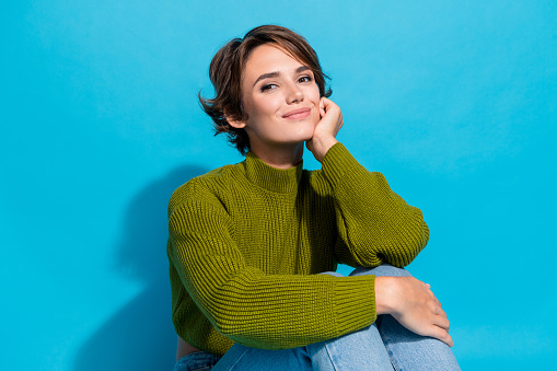 Photo of charming dreamy cute girl wear green winter clothes sit floor arm touch cheeks isolated over cyan color background.