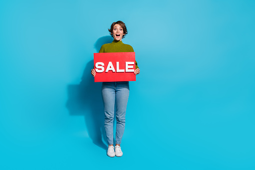 Full length photo of amazed cheerful lovely girl wear stylish outfit hold card presents sale empty space isolated on cyan color background.