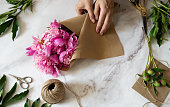 Florist at work: pretty woman making summer bouquet of peonies on a working gray table. Kraft paper, scissors, envelope for congratulations on the table. View from above. Flat lay composition.