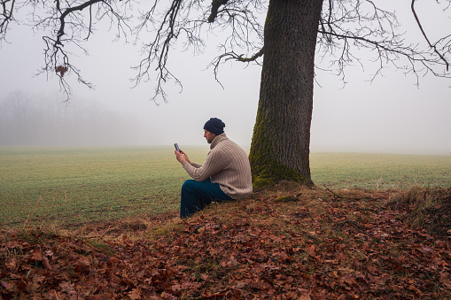 Young man in sweater sitting near tree using mobile phone in morning fog. Czech landscape, relax concept