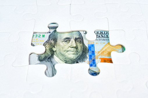 One hundred dollar bill elements seen through the openings in a jigsaw puzzle background. Financial aspirations, opportunity and strategic thinking related concept.