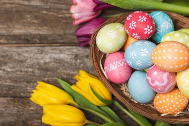Easter basket filled with colorful eggs and a bouquet of tulips on  table. Easter celebration concept. Colorful easter handmade decorated Easter eggs. Place for text. Copy space