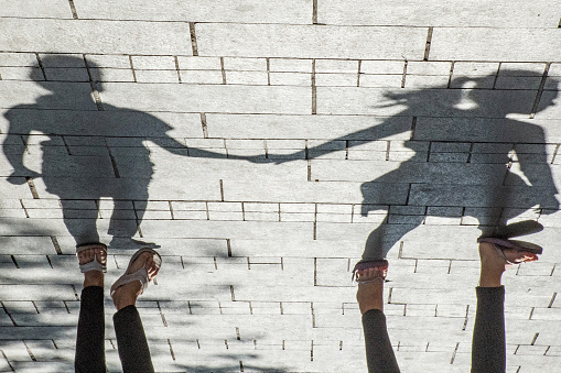 Feet and sandals and the shadow silhouette of two young girls holding hands cast on a sunlit brick sidewalk.