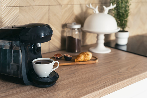 Selective focus on coffee maker machine and cup with fresh drink on wooden countertop at modern kitchen in morning. Hot espresso and croissant prepared for breakfast at home.