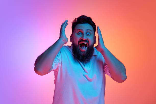 istock Surprised man widely open eyes and mouth looking at camera raising hands in neon light against vibrant gradient studio background. 2048168053