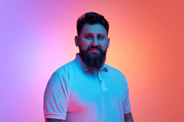 istock Calm bearded man looking at camera in neon light against vibrant gradient studio background. Male model looks confident. 2048168058