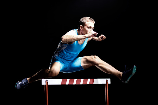 Mid-air shoot of a young man jumping over hurdle in a track race. Stadium light in the back.