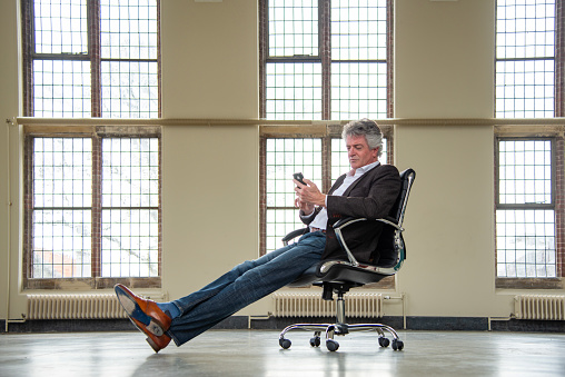 Mature businessman sitting on office chair, waiting, looking at smartphone. Setting is a large empty hall