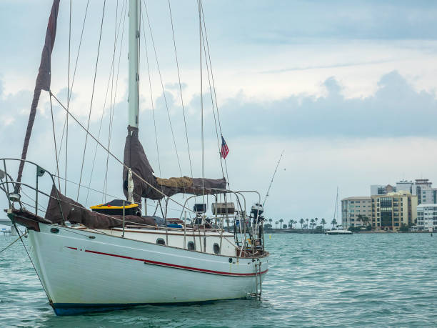 Sailboat moored in Sarasota Bay, its mainsail wrapped around the boom, with partial view of city skyline and approach to causeway, on an overcast afternoon in Florida. For nautical and travel motifs.