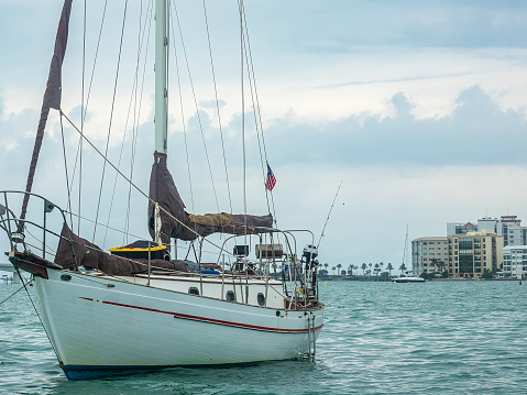 Sailboat moored in Sarasota Bay, its mainsail wrapped around the boom, with partial view of city skyline and approach to causeway, on an overcast afternoon in Florida. For nautical and travel motifs.