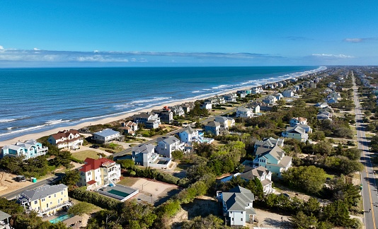 From high above, Duck, North Carolina presents a breathtaking panorama. The sun casts a warm glow over the beach where the blue waves gently lap against the golden sands. A row of beachfront homes, each painted in cheerful hues, add a splash of color to the natural beauty of the Outer Banks. The emerald-green vegetation offers a stark contrast to the sandy shore, completing this picturesque seaside tableau.