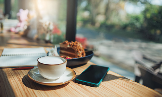 Hot fresh milk in coffee cup with chocolate cake for breakfast and mobile, note book on wooden table and outdoor garden background