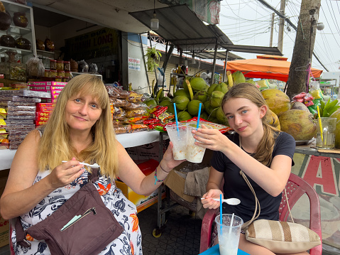 Two family member tourists eat sticky coconut, from a road side street vendor. this retail space displays coconuts and various sweets made from this  traditional  crop. colourful shop canopies are in the background.