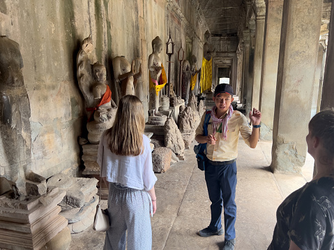 Two tourists are standing listening to a guide talking about a feature at the Angkor Wat site. The family of European tourists are situated , along an open avenue of architectural columns and many Hindu /Buddist stone figures. Several of the stone statues are partially robed in swathes of saffron and orange fabric.