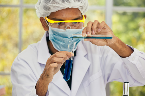 Close-up of a meticulous chemist in a lab coat and safety glasses pouring a blue chemical substance between test tubes against a bright window