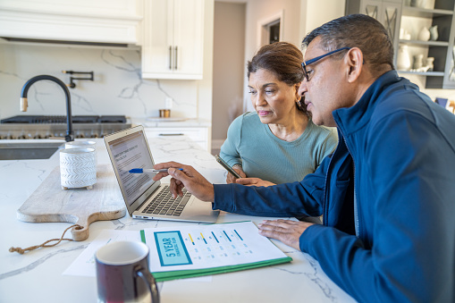 A couple sit at their kitchen island with a laptop open and their bills spread out in front of them as they review their finances together.  They are dressed casually and appear inquisitive.