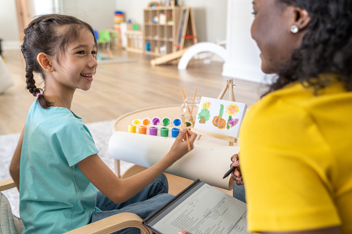A female therapist of African decent, sits at a table with a little girl during an art therapy session.  The little girl has a small canvas on an easel and colourful paints out, and the therapist is taking notes electronically.