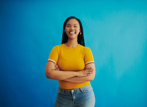 A young black woman wearing a yellow t-shirt exuding confidence with crossed arms and a bright smile, symbolising empowerment and self-assurance, shot against a blue background