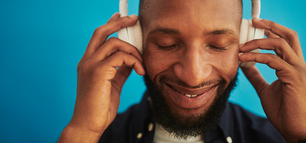 A happy young African man enjoying music with headphones, shot against a blue background with copy space. Stock photo