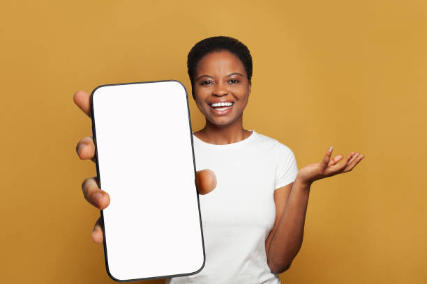 istock Joyful woman showing smartphone with empty blank screen display. Model in white t-shirt with phone on yellow studio wall background 2048445875