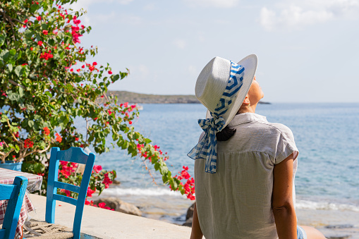 Unrecognizable woman with Greek hat enjoying a splendid morning by the sea and next to a typically Greek tavern with flowering plant