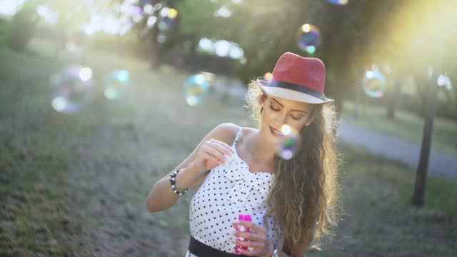 Young Woman In A Polka Dot Sundress Wearing A Hat Blowing Soap Bubbles In A Sunny Park