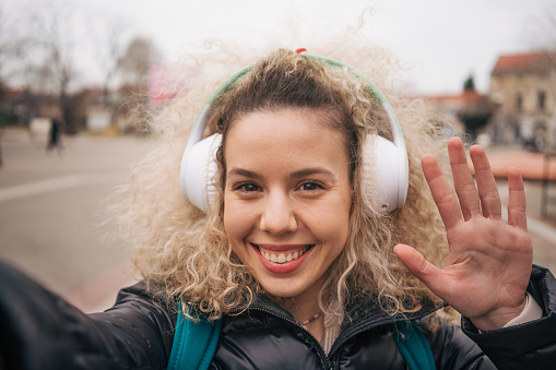 Portrait of a young curly woman holding a headset on her head and talking to someone on a video call. She waves and looks at the camera.