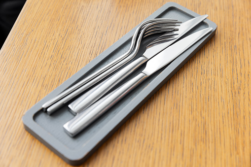 Shiny forks and knives made of stainless steel lay on small gray wooden tray on an empty  wooden table