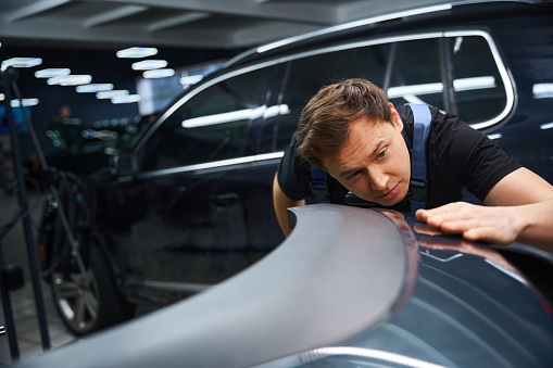 Serious focused collision repair technician inspecting surface of car fender after dent removal