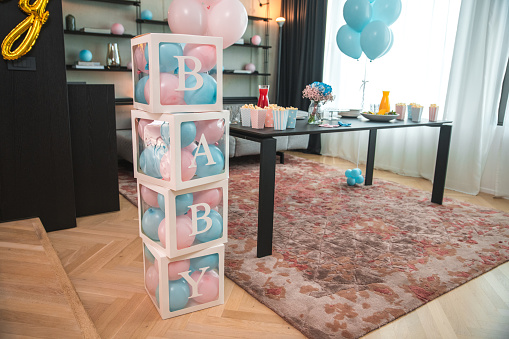 A festive indoor gender reveal party scene featuring large alphabet blocks spelling 'BABY', adorned in blue, signaling the upcoming arrival of a boy. The decor includes blue balloons and a table set with snacks and drinks, creating a celebratory atmosphere in a stylish home setting.