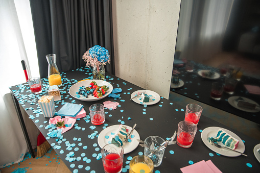 Celebratory scene of a gender reveal party indoors with blue-themed table setting featuring confetti, sweets, and beverages, indicating the expected baby is a male. Casual party attire and a lively domestic setting are evident.
