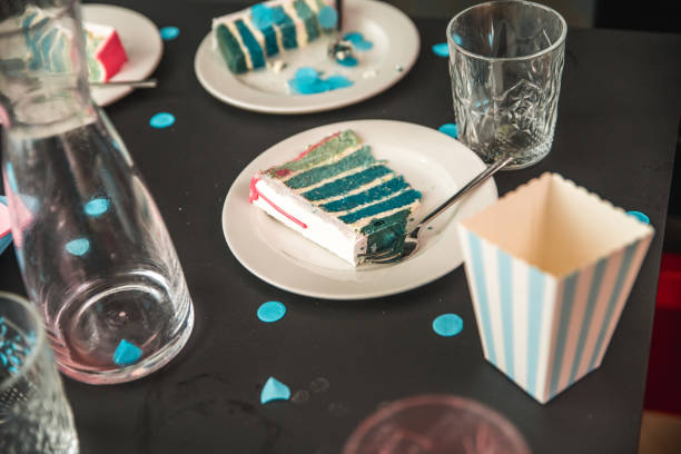 Gender reveal party scene with a slice of blue layered cake on a plate indicating a boy, surrounded by themed decorations and confetti. Festive table with casual utensils and a hint of the celebratory event indoors.