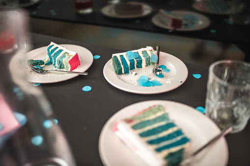Close-up view of white plates holding slices of blue layered cake, indicating the reveal of a baby boy at a gender reveal party. The table setting features casual dining with scattered blue confetti, signaling a festive occasion indoors.