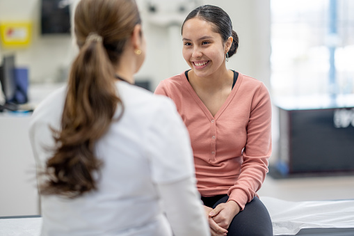A young female of Hispanic decent, sits up on an exam table as she meets with a doctor to discuss her health concern.  She is dressed casually and is smiling at her doctor who is seated in front of her.
