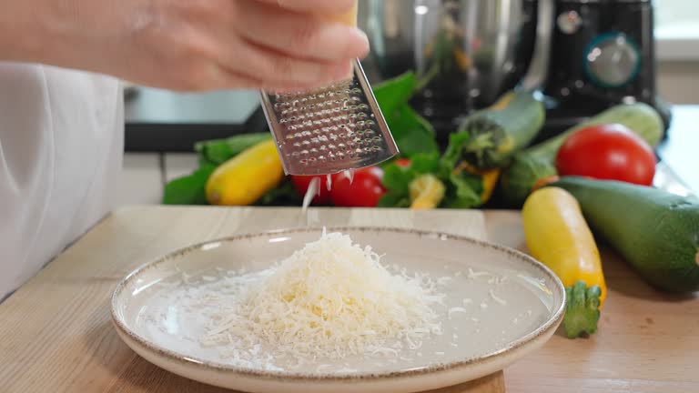 Close-up of a chef's hands grating parmesan cheese onto a fine grater into a plate, wooden table, vegetables on the background, front view