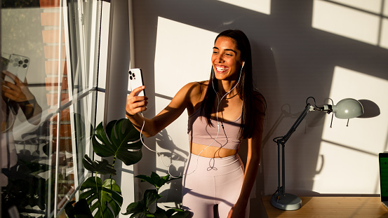 Portrait of an beautiful young woman of Brazilian/Hispanic ethnicity, listening to music on headphones and taking selfie with mobile phone, near the window during sunny day