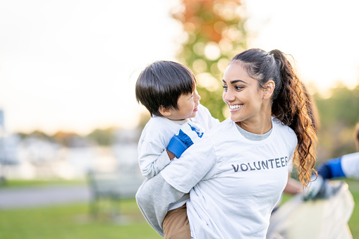 A young woman gives a little boy a piggy-back ride as they have fun during a community volunteer event.  They are both wearing matching white Volunteer t-shirts and laughing.