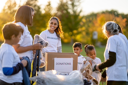 A small group of volunteers gather together in a park to sort local donations and help their community. They are each dressed casually and are wearing matching white Volunteer t-shirts.