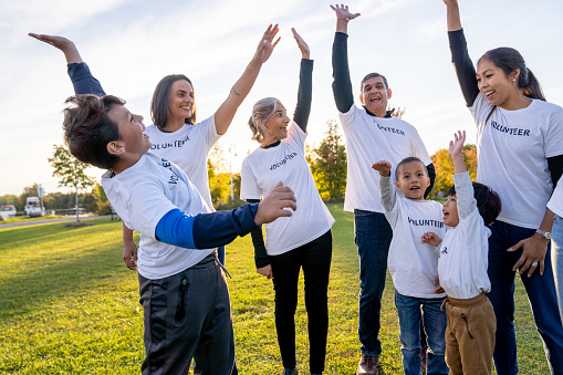 A small group of local volunteers gather in a circle as they throw up their hands and cheer each other on to begin their work.  They are each dressed casually and are wearing matching white Volunteer t-shirts.