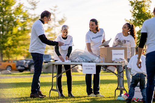 A small group of volunteers gather together in a park to sort local donations and help their community. They are each dressed casually and are wearing matching white Volunteer t-shirts.