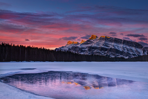 Golden sunrise at Two Jack Lake with alpenglow on Mount Rundle peaks reflecting off the frozen lake covered with snow and ice.