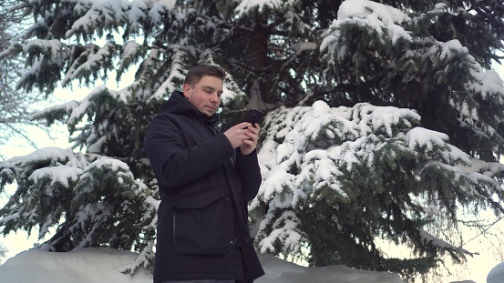 A young man stands on the phone against a background of spruce in winter. A man with a smartphone chats against the backdrop of snowy spruce branches. 4k