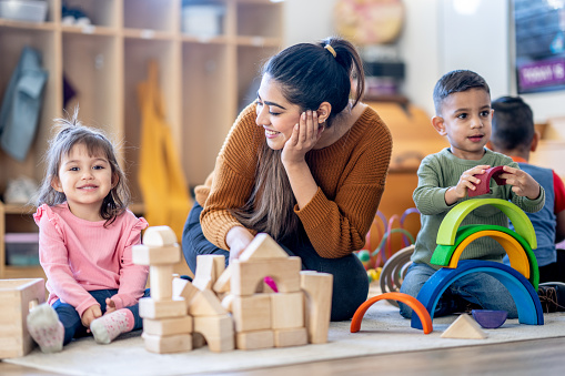 A female elementary teacher of Middle Eastern decent, sits on the floor with a little girl as they build together with wooden blocks.  Other students can be seen playing individually in the background.