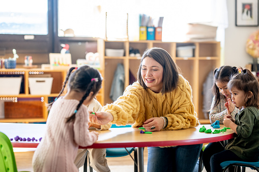 A small group of elementary students sit with their teacher around a table as they play together with Play-Doh.  They are each dressed comfortably and are focused on their individual creations.