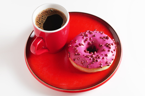Pink donut with sugar balls and cup of coffee on red utensils on white background