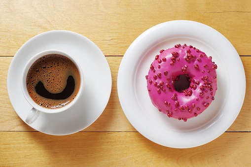 Pink donut with red sugar balls sprinkles and cup of coffee on wooden table, top view