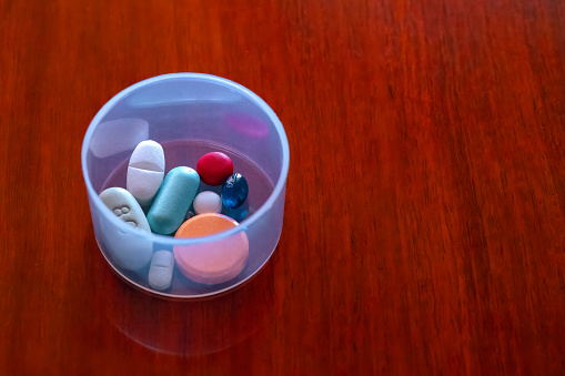 A cocktail of vitamins and medicines for the morning in a plastic container, on a rich shiny rosewood table. Shot in ambient morning light. Copy space.